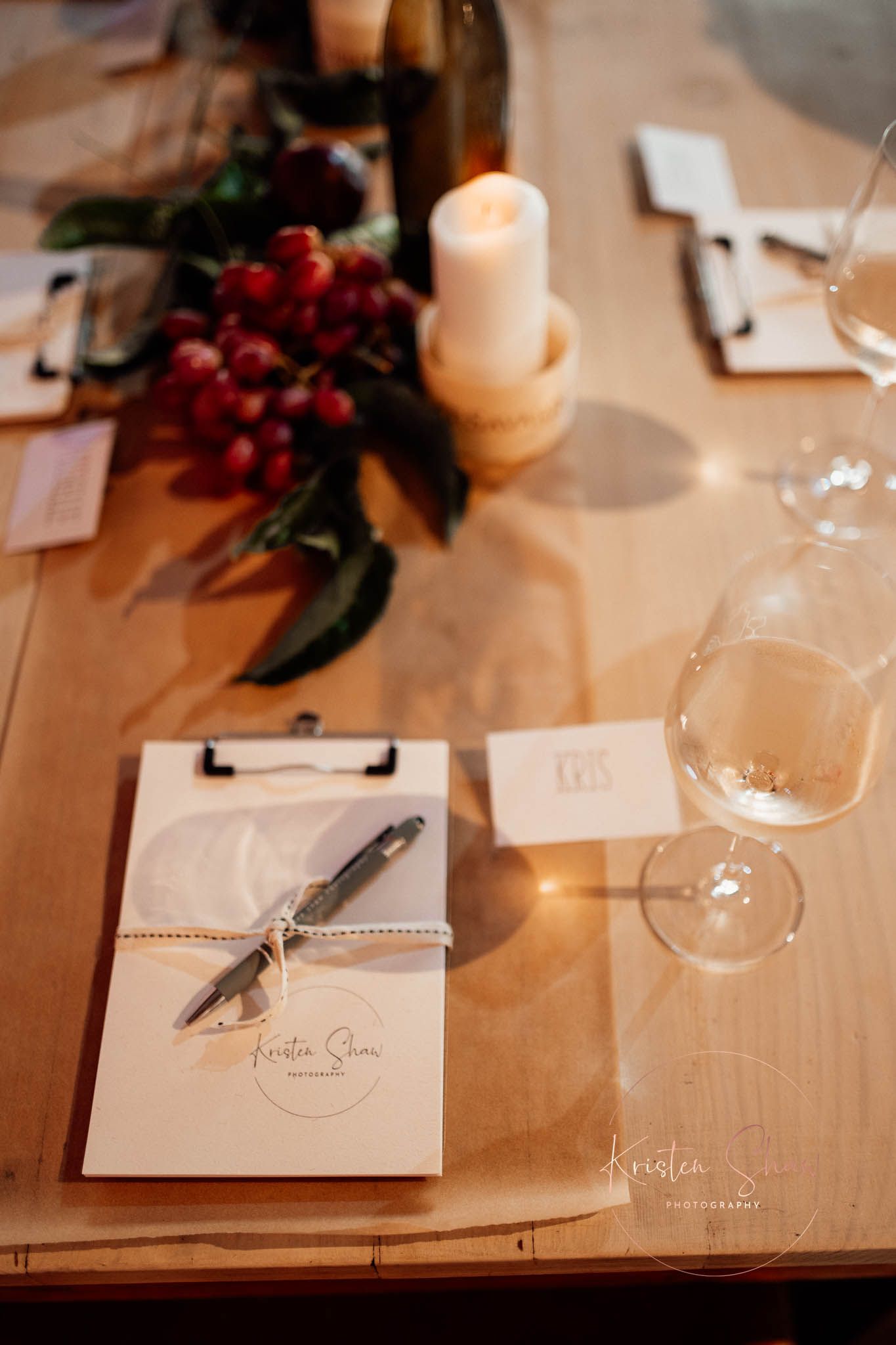Elegant wedding table setting with clipboard, pen, wine glasses, candle, and red berry centerpiece on wood table.