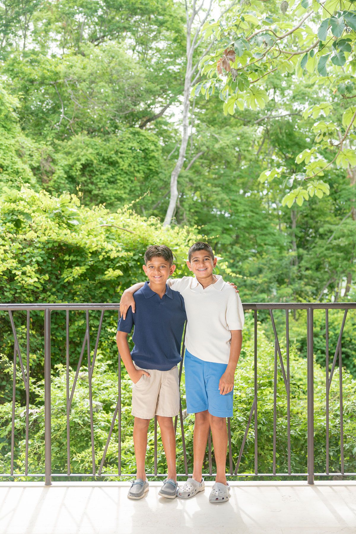 Two smiling boys standing together on a deck with lush green trees in the background.