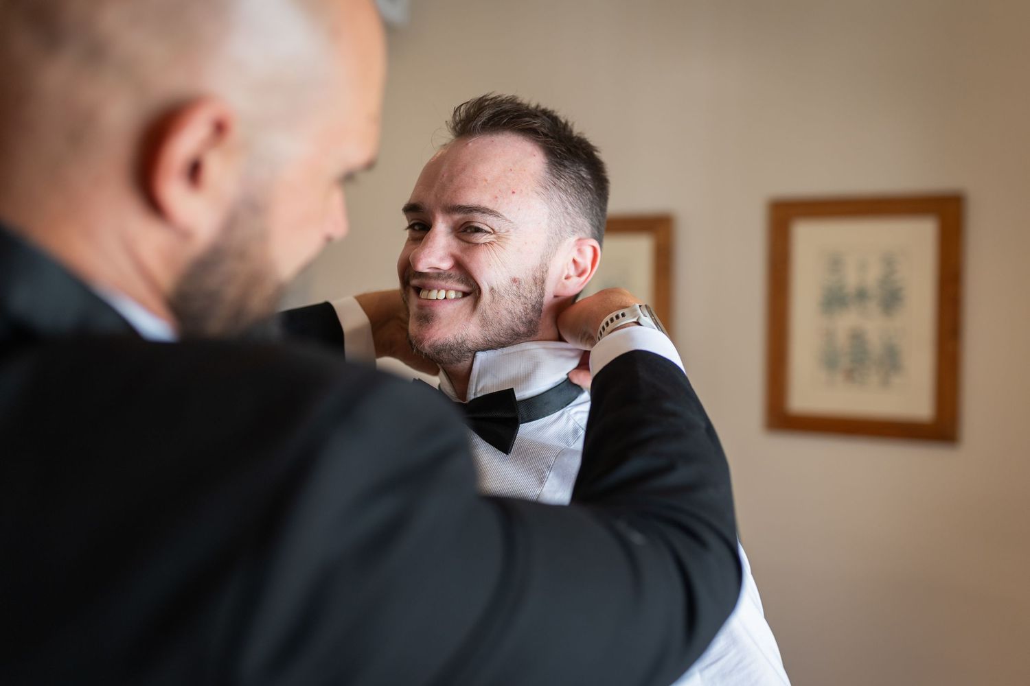 Groom smiling as best man adjusts his bow tie before wedding ceremony in elegant indoor setting.