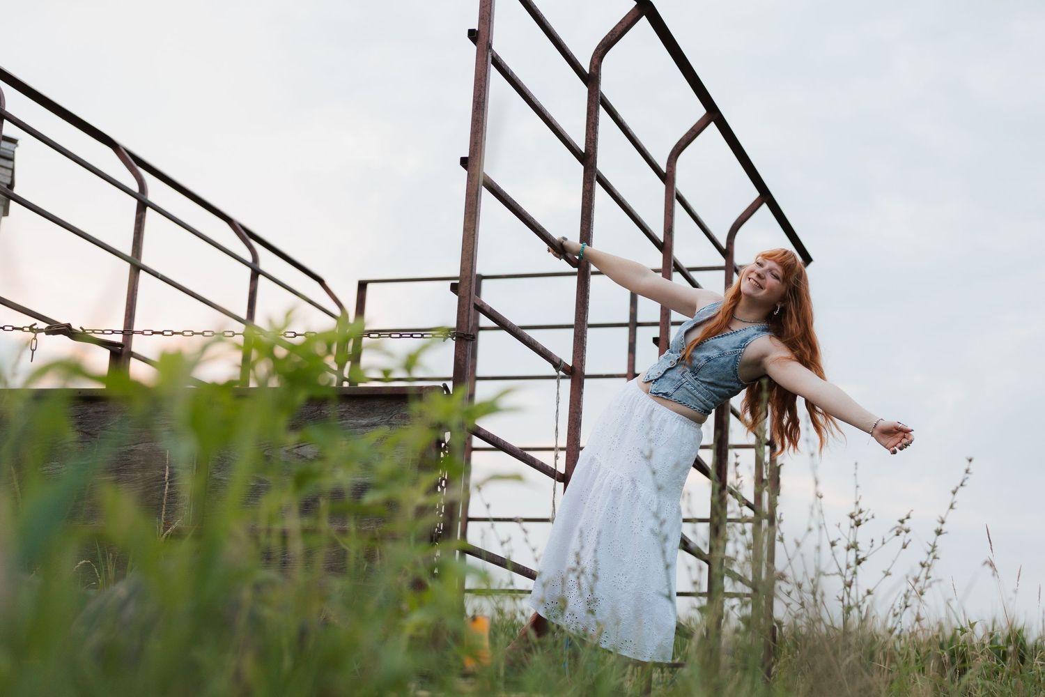 Young woman in white skirt and denim top posing joyfully outdoors near rustic metal stairs in a green field.