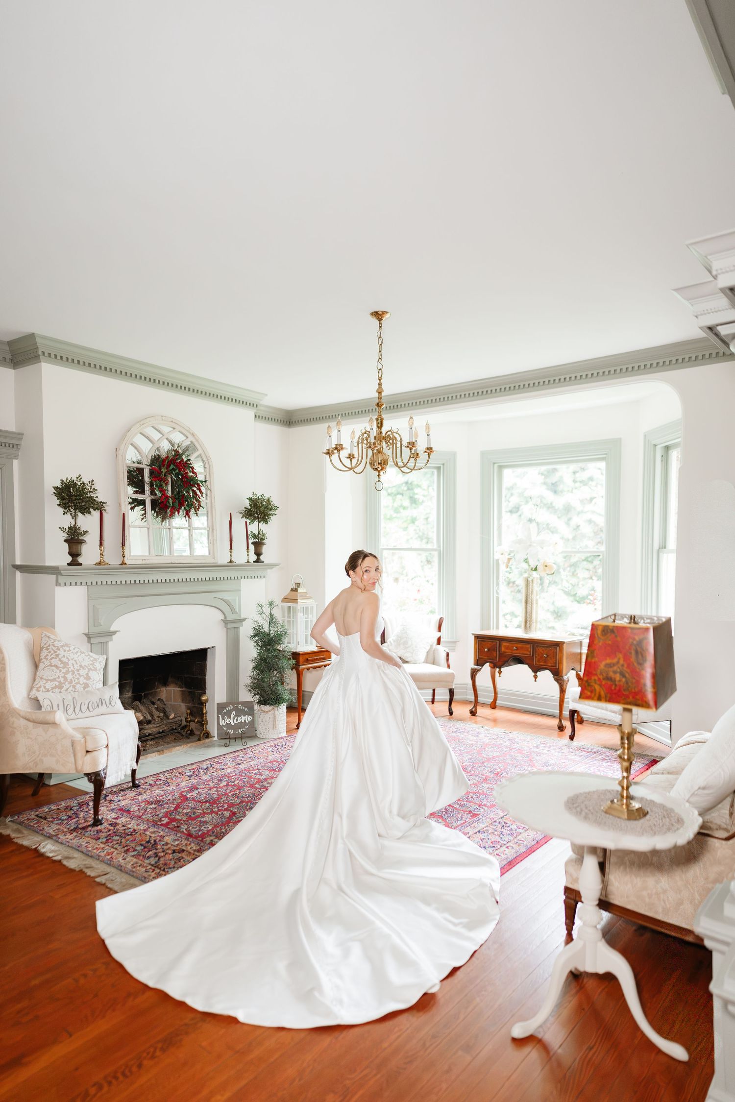 Bride in backless white ball gown with long train stands in elegant room with fireplace and chandelier.