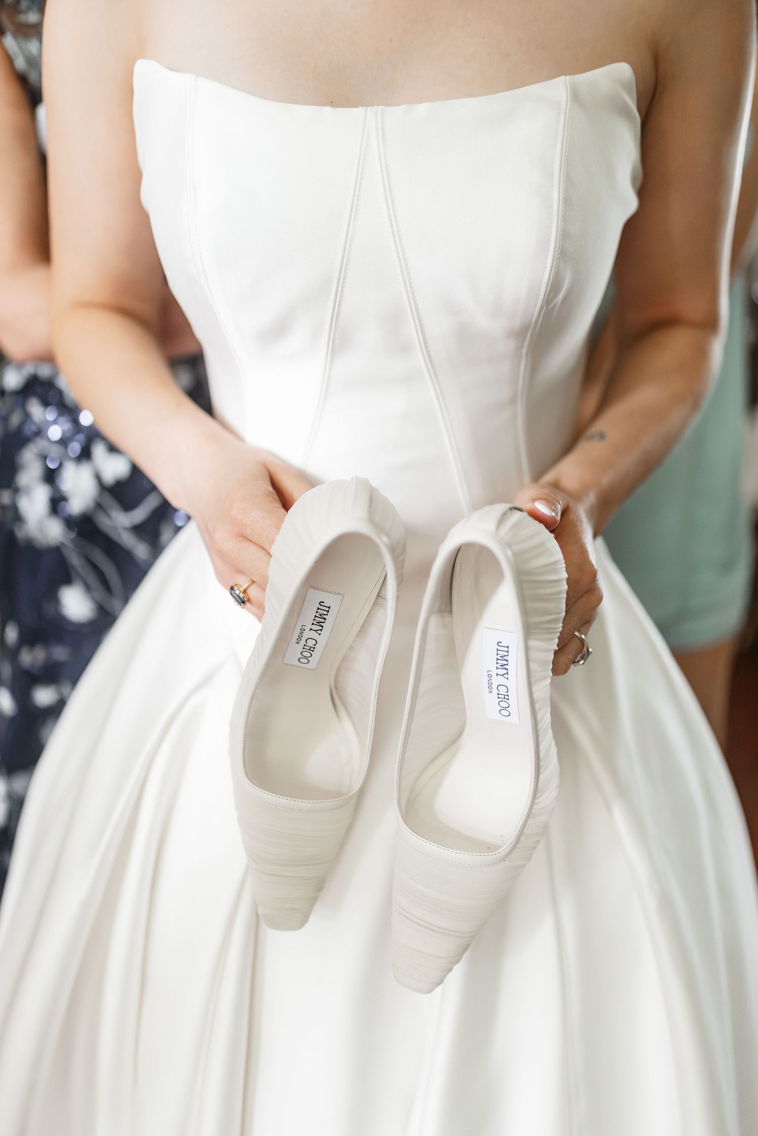Bride in strapless white wedding dress holds up Jimmy Choo ivory bridal shoes toward camera.