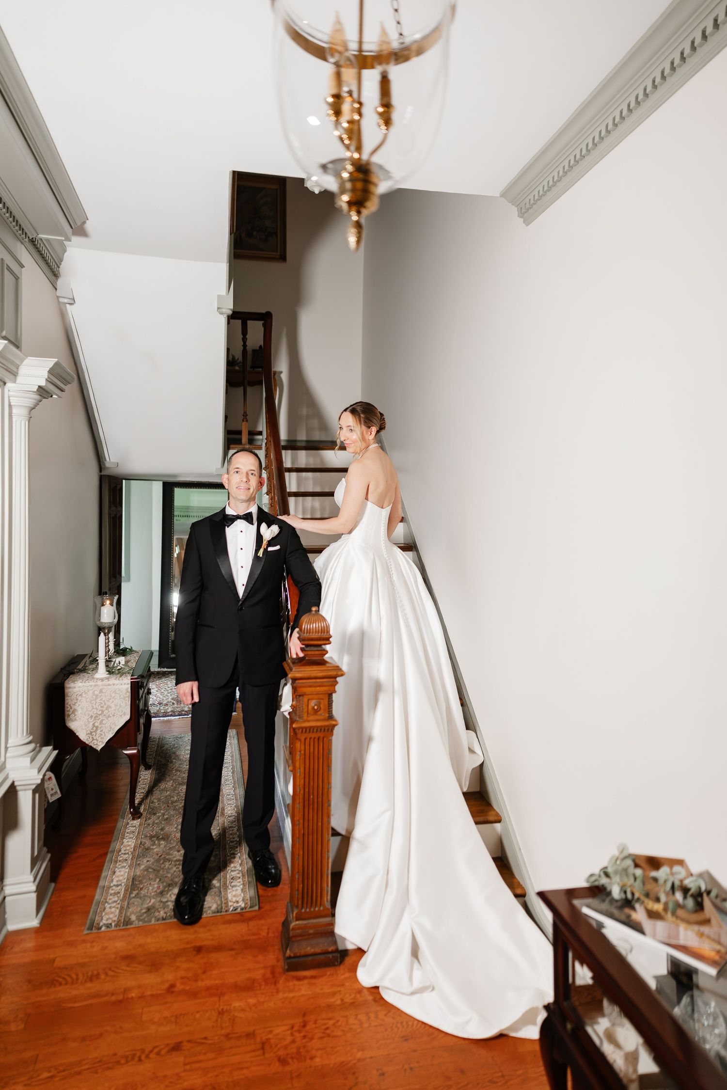 Bride in a strapless white gown descends a staircase as groom in black tuxedo waits in a classic foyer.