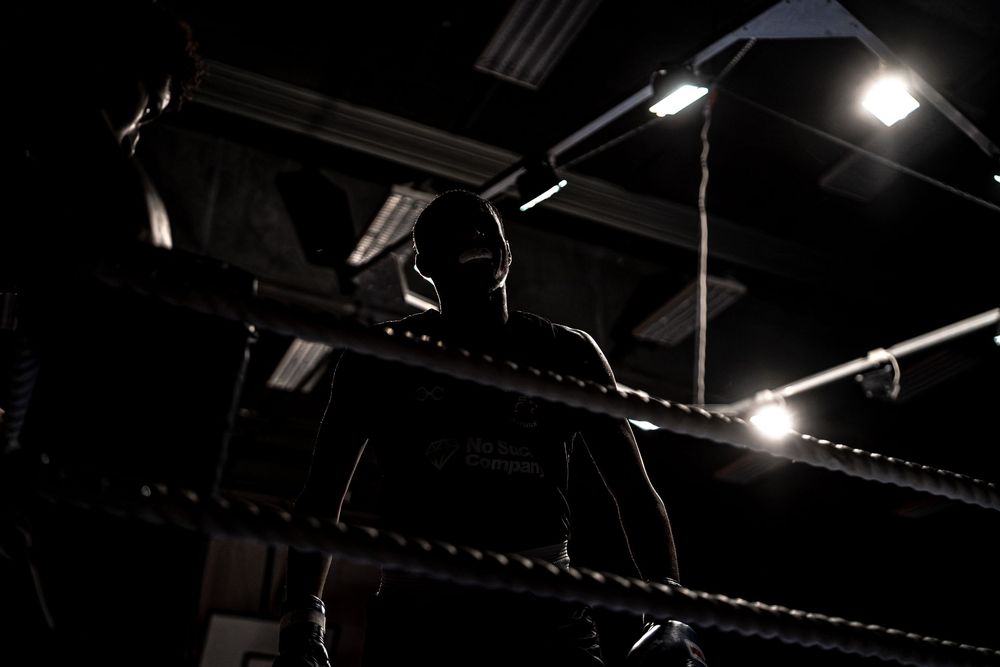 Silhouetted boxer standing in a dark boxing ring gym, dramatic low-light atmosphere with bright overhead light.