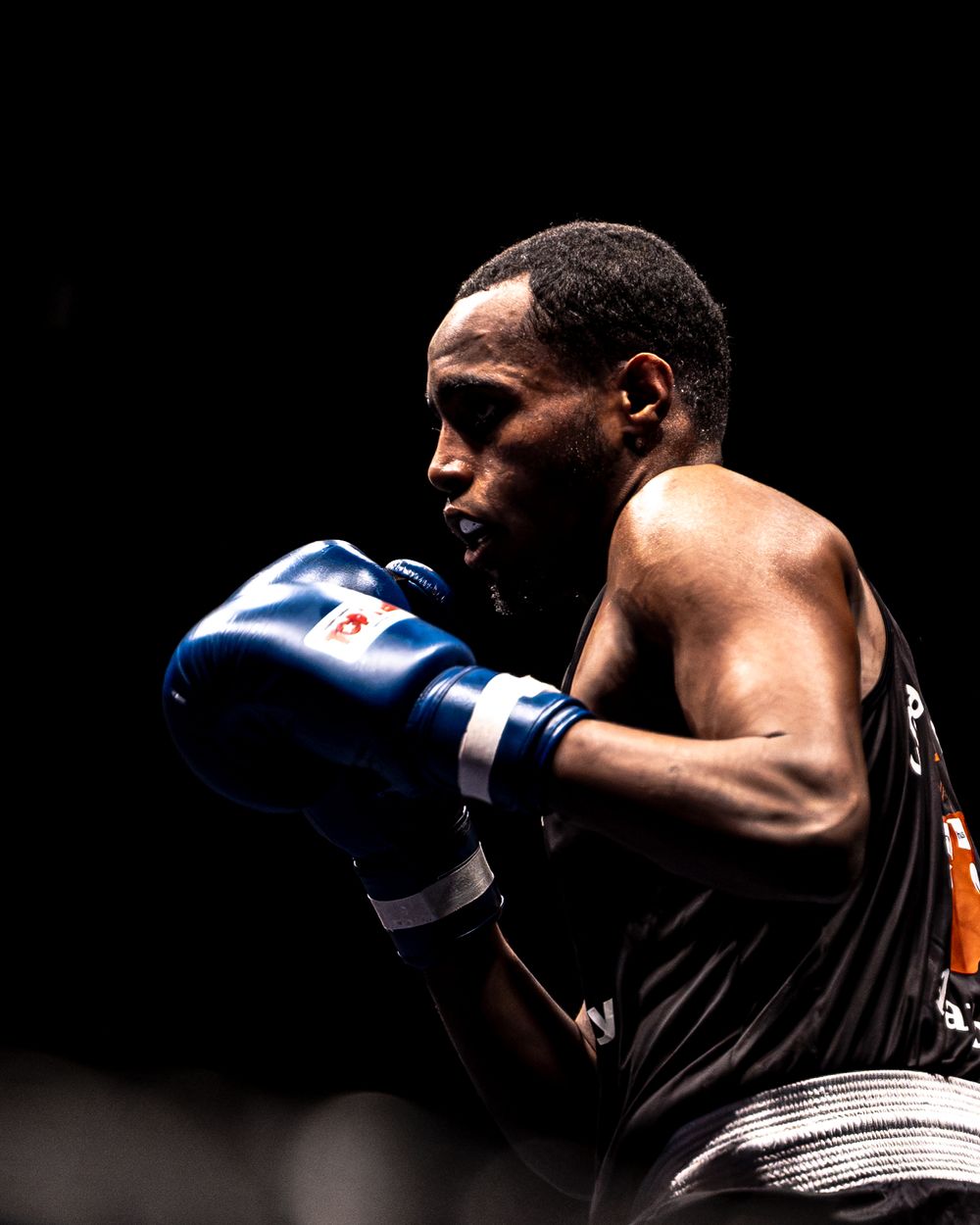 A focused boxer wearing blue gloves in a fighting stance against a dark background.