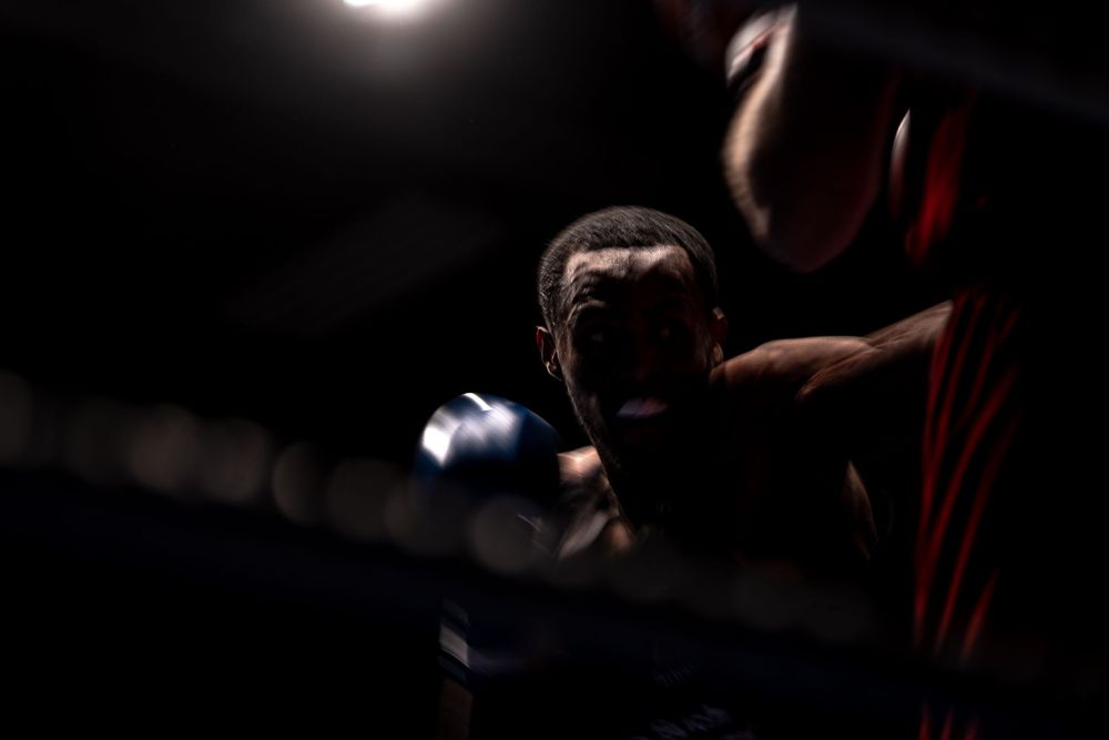 A boxer in blue gloves stands in a dimly lit boxing ring, illuminated by dramatic overhead lighting.