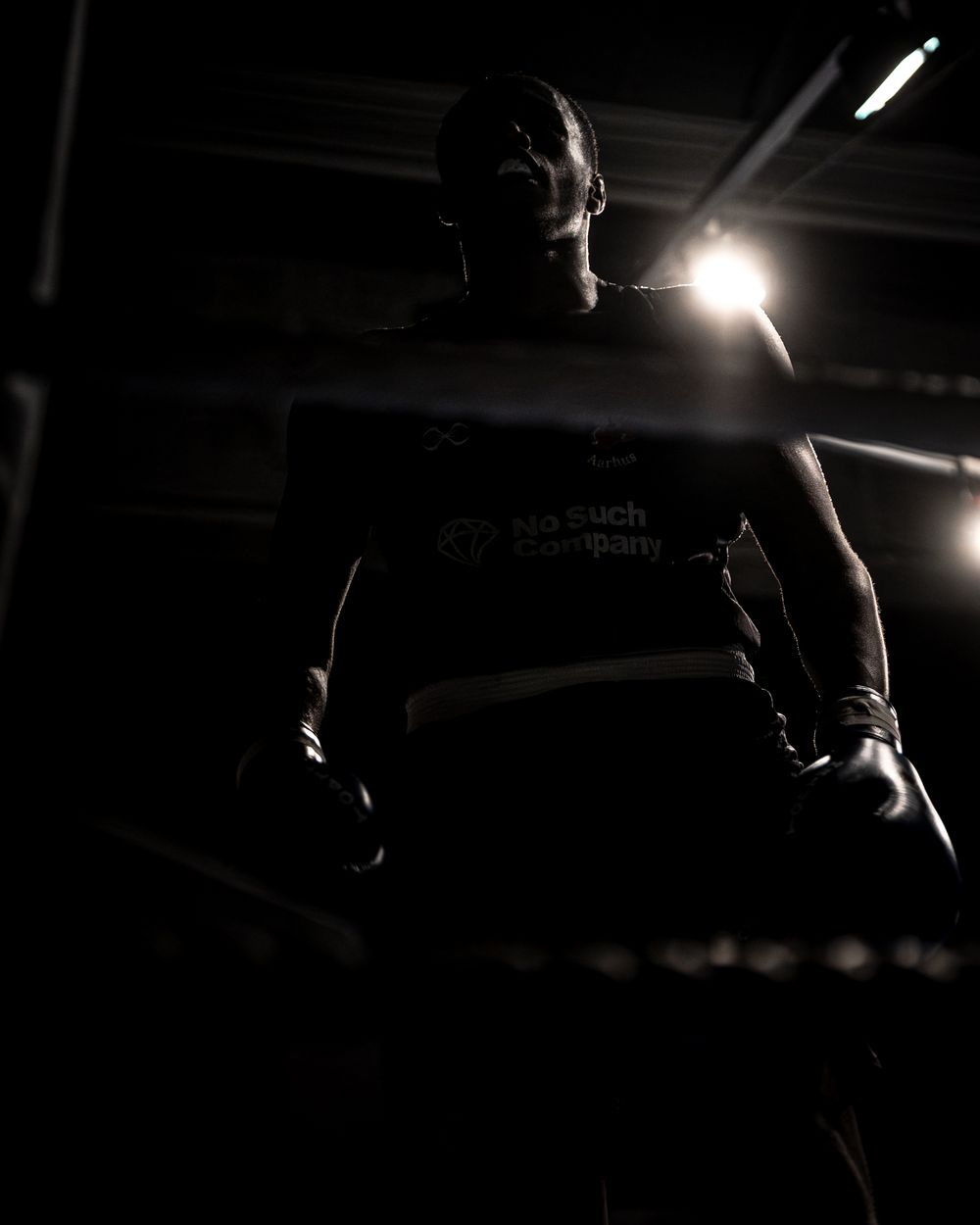 Silhouetted boxer standing in a dark gym with dramatic backlighting highlighting their athletic frame.