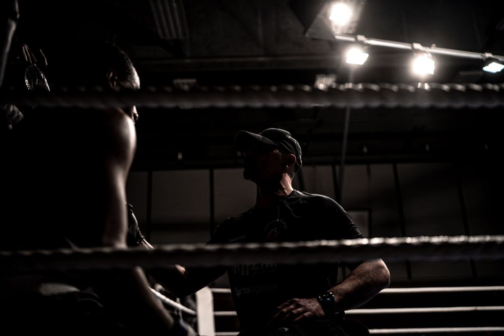 A boxing trainer watches attentively from the corner of a dimly lit boxing ring during a match.