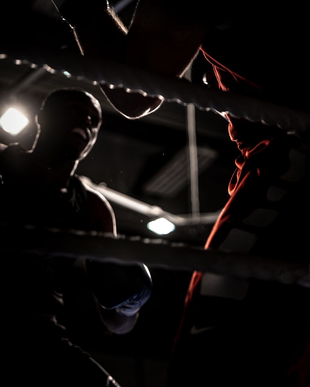 Two boxers face off in a dark boxing ring, illuminated by dramatic lighting through the ropes.