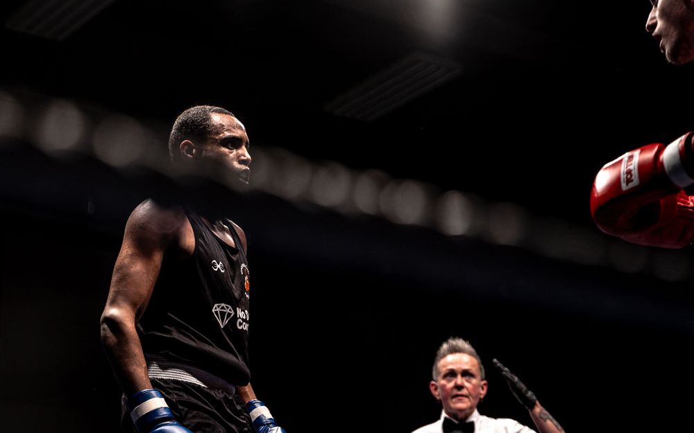 A muscular boxer in black attire squares up against an opponent while a referee watches closely.