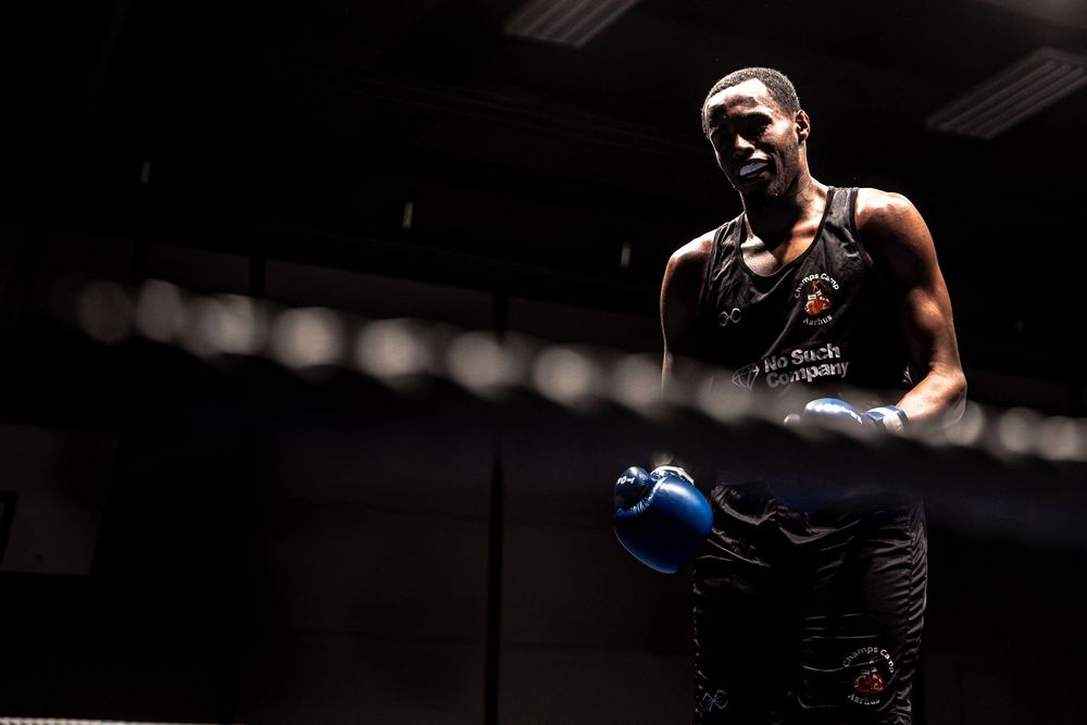 A boxer in dark gym wearing blue gloves and black shorts stands confidently under dramatic lighting.