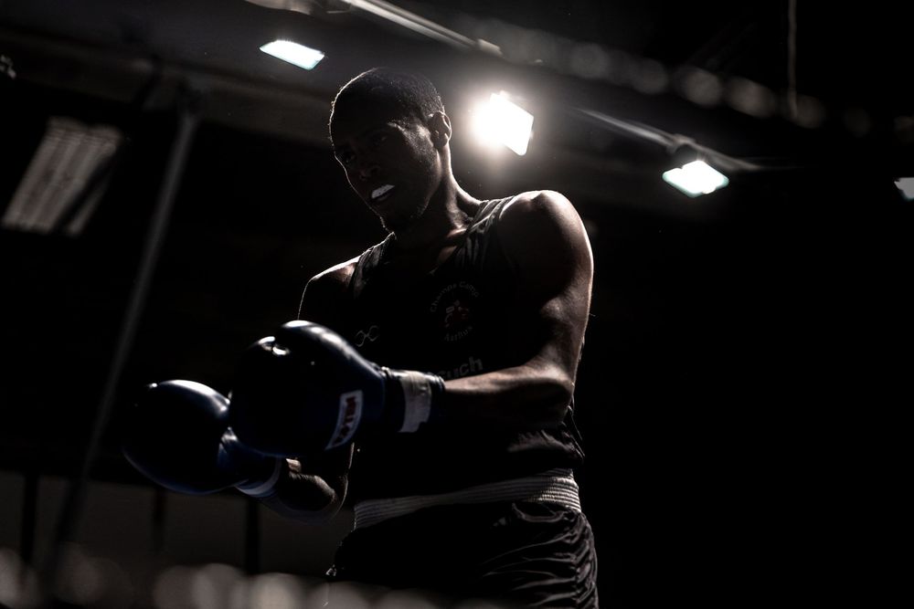 A male boxer in dark gym lighting stands ready with blue boxing gloves under a bright spotlight.