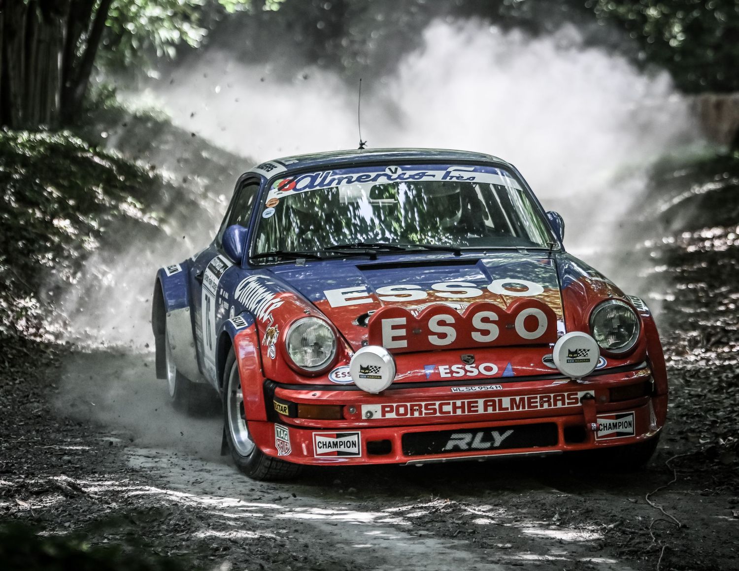Red Esso-sponsored Porsche 911 rally car racing on a dusty forest track, kicking up dirt and debris.