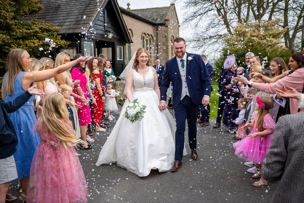 Bride and groom walk through confetti shower with smiling guests lining the path outside a stone wedding venue.