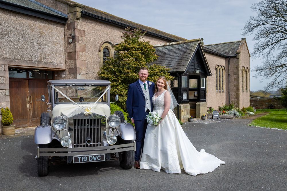 Bride and groom posing beside a vintage silver wedding car outside a rustic stone church venue.