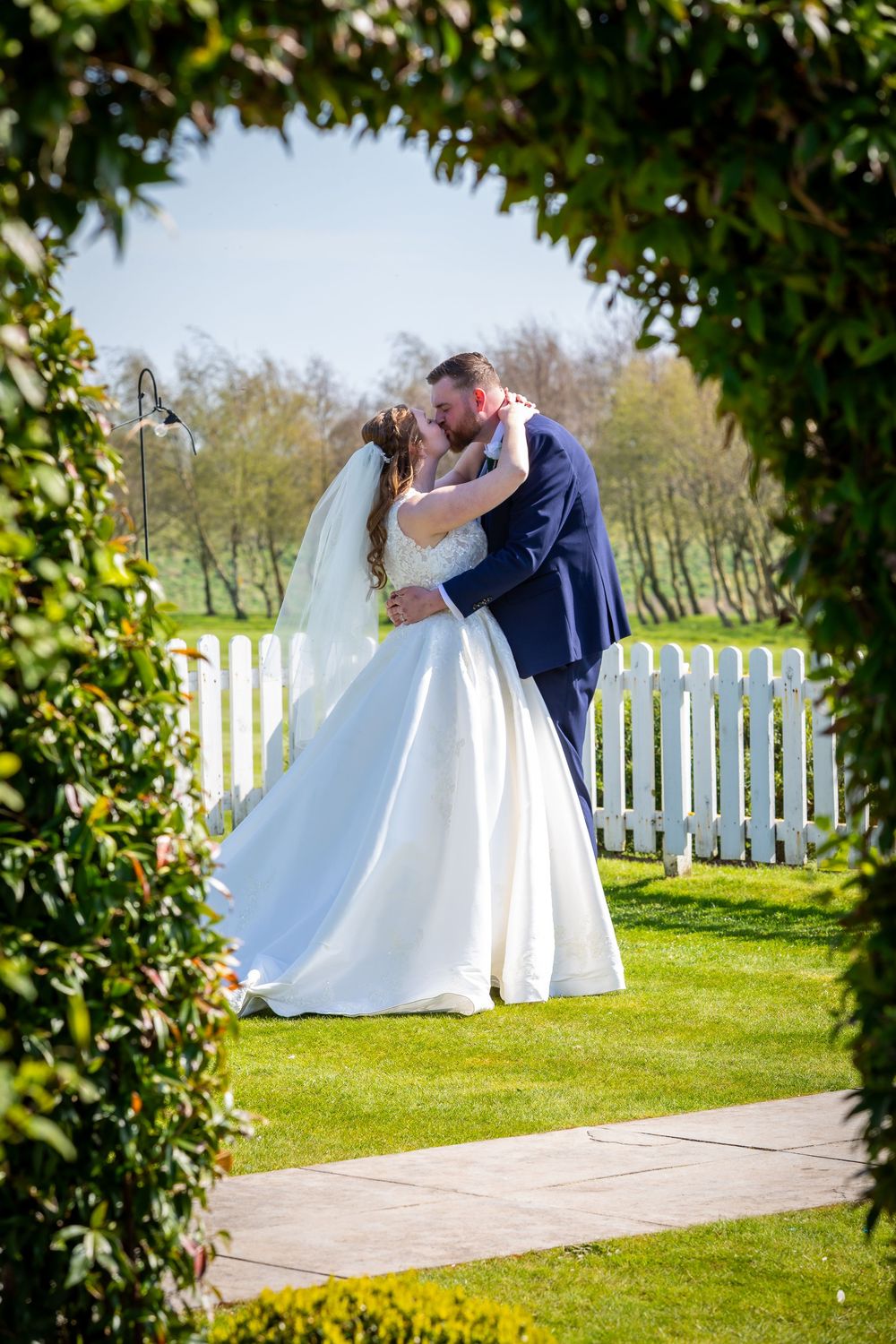 Bride and groom share a romantic kiss framed by a lush green hedge arch on a sunny wedding day.