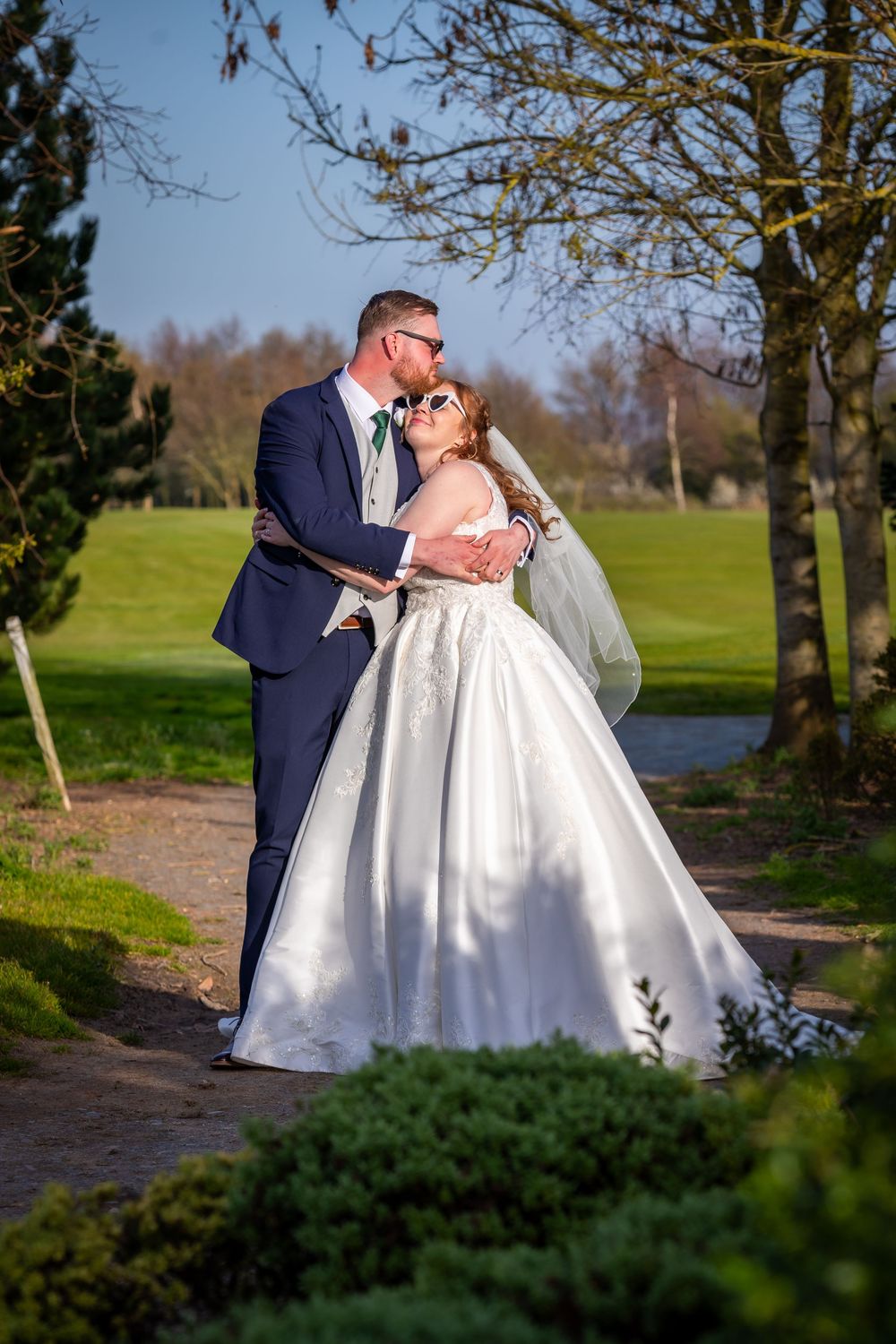 Bride and groom share a romantic kiss outdoors on their wedding day, surrounded by lush green trees and gardens.