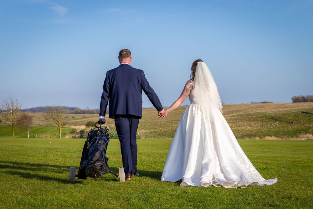 Bride and groom holding hands walking on green golf course grass under blue sky on their wedding day.