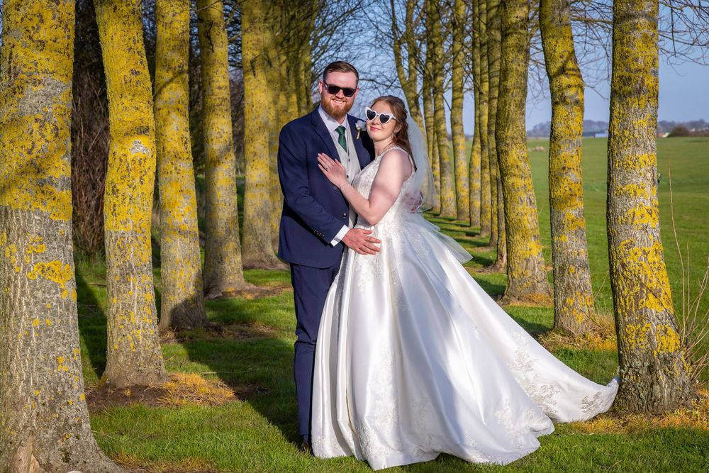 Bride and groom pose elegantly among tree-lined avenue on lush green lawn in stunning outdoor wedding portrait.