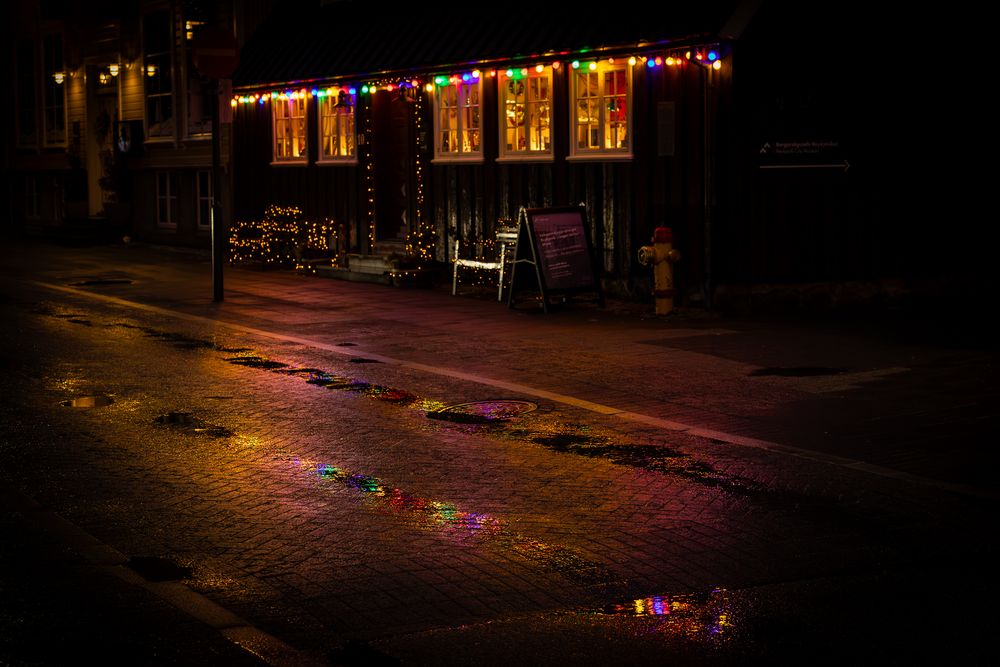 Colorful Christmas lights reflecting on wet cobblestone street outside a festively decorated shop on a rainy night.