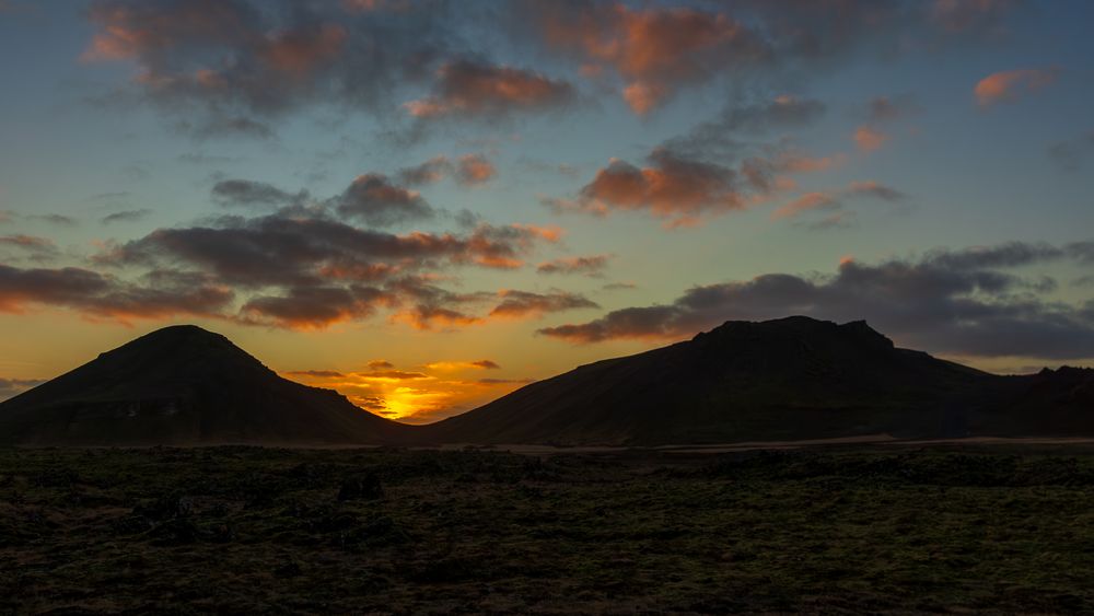 Dramatic sunset behind two volcanic hills with colorful clouds illuminated in orange and purple.
