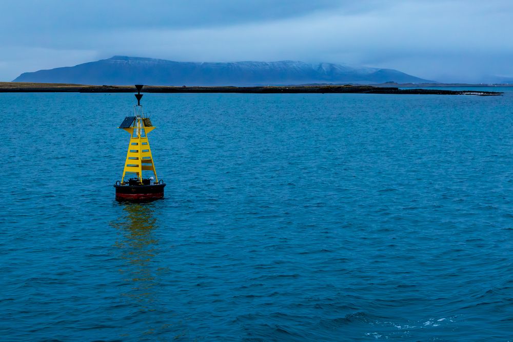Yellow navigational buoy floating in calm blue harbor waters with a snow-dusted mountain island visible in the background.
