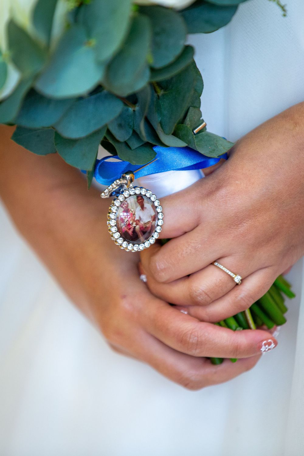 Bride holding wedding bouquet with blue ribbon and diamond-framed memorial photo charm, wearing engagement ring.