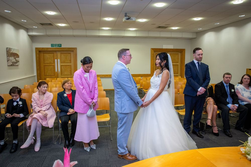 Couple exchanging vows at a civil wedding ceremony in a modern register office with seated guests.