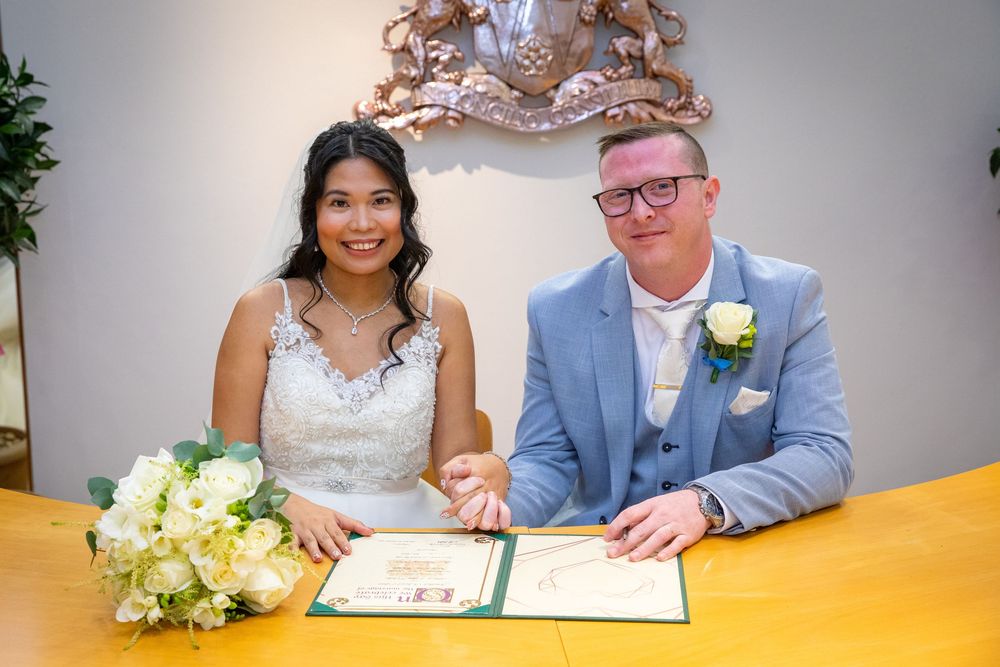 Bride and groom smiling while signing wedding documents at a registry office table with a bridal bouquet.