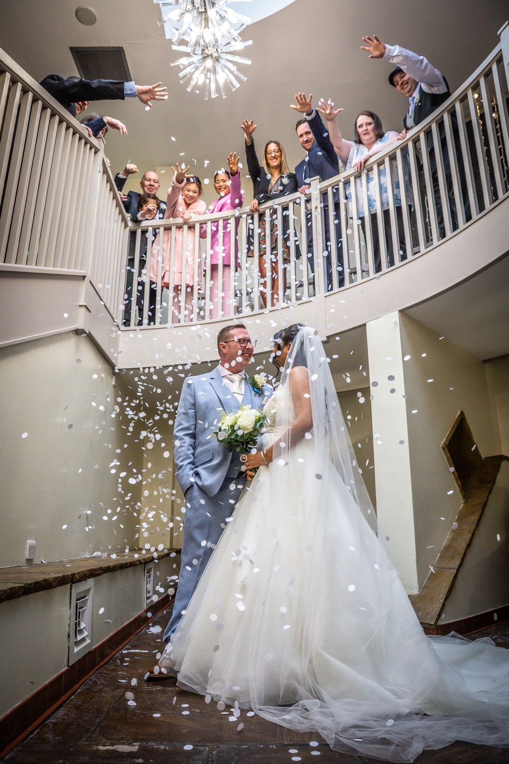 Bride and groom share a romantic moment as confetti falls, surrounded by cheering guests on a curved staircase.
