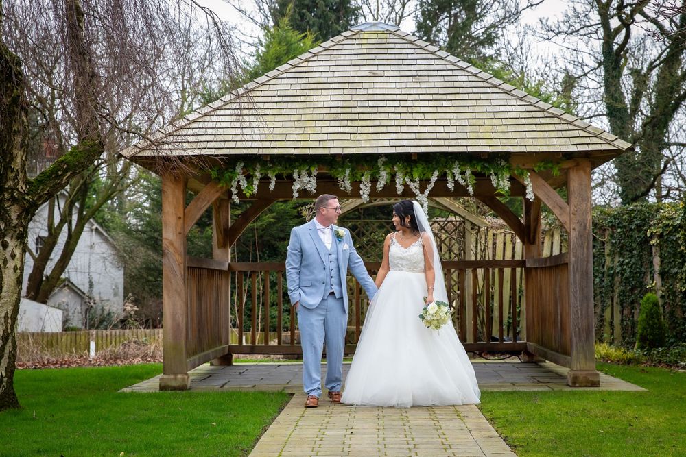 Bride and groom holding hands in front of a wooden gazebo decorated with flowers in a lush garden setting.