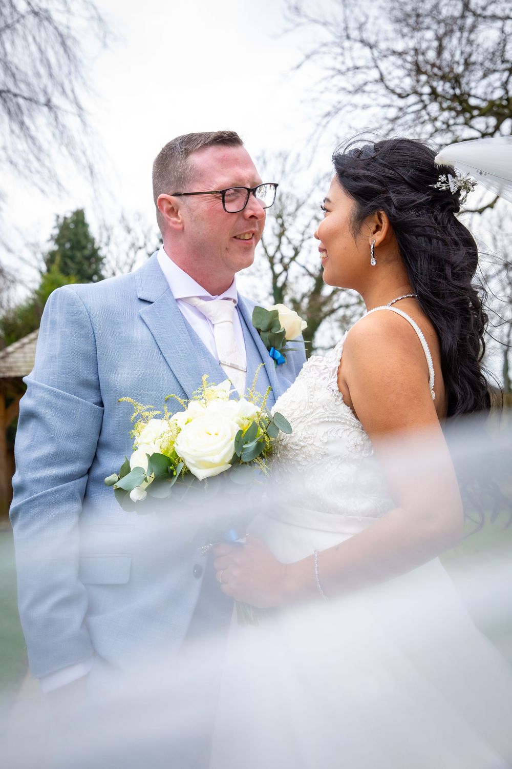 Bride and groom gazing at each other outdoors, she holds white roses, he wears a light blue suit, veil softly blurs foreground.