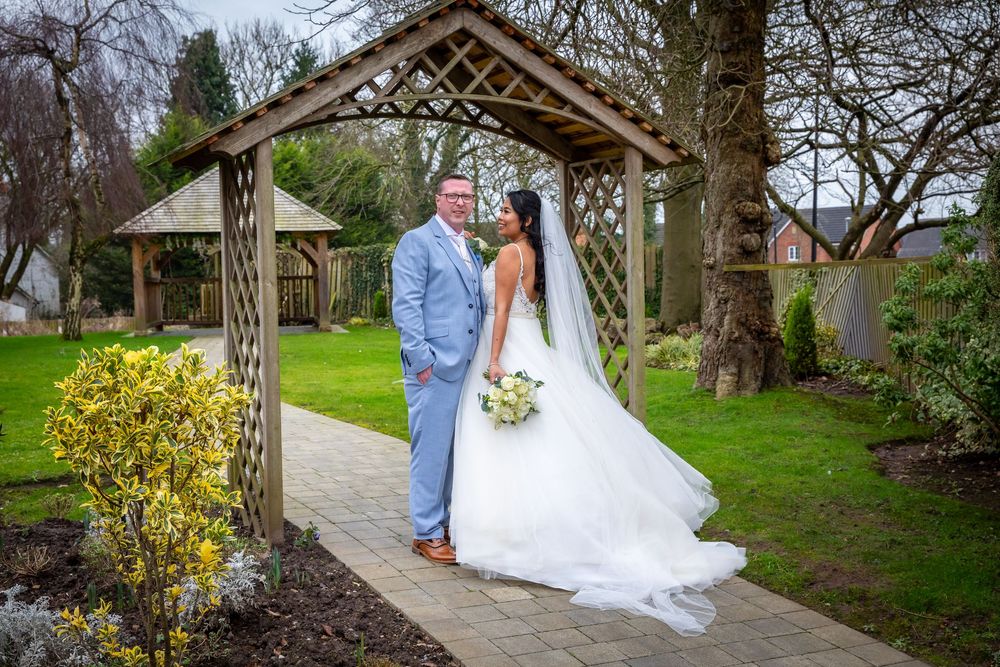 Bride and groom pose under a wooden pergola in a lush garden on their wedding day.