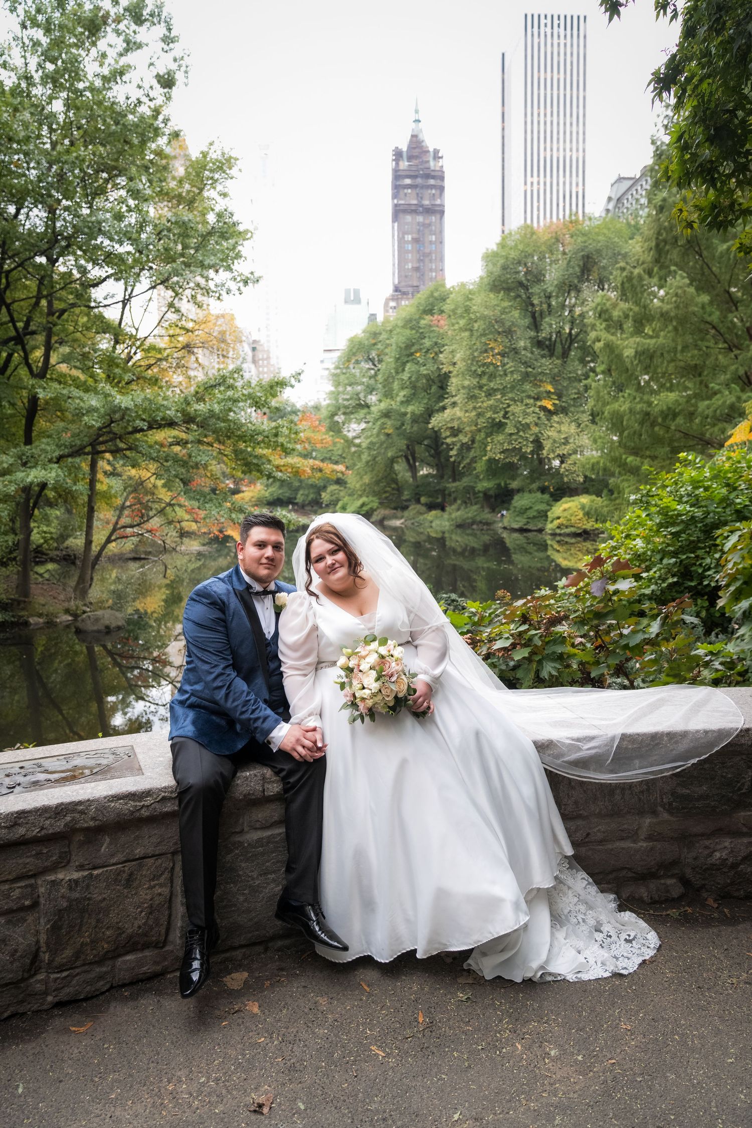 Bride and groom pose together on a stone bridge in Central Park, NYC, with lush trees and skyline in the background.