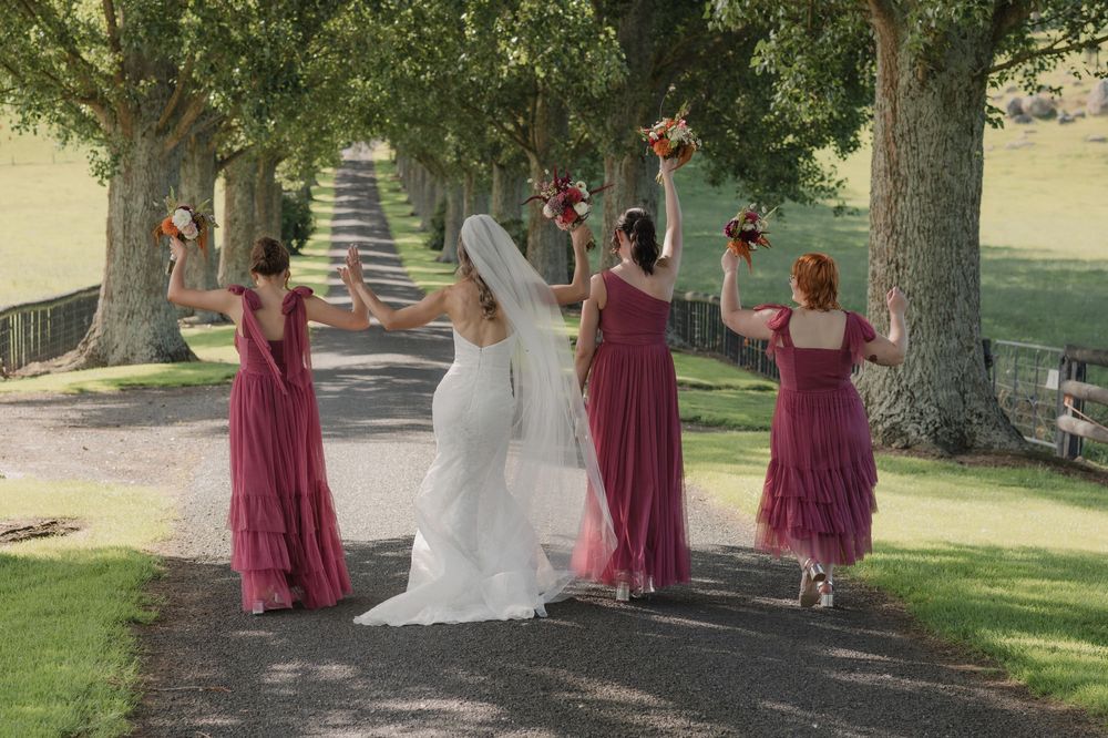 Bride in white gown and bridesmaids in burgundy dresses walk away raising bouquets under tree-lined path.