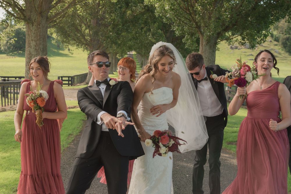 Wedding party poses playfully on a tree-lined path, bride in white gown with bridesmaids in dusty rose dresses.