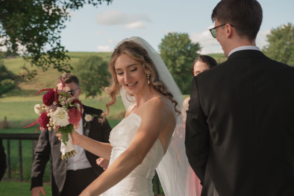Bride in white gown holding floral bouquet smiles during outdoor wedding ceremony with lush green hills backdrop.