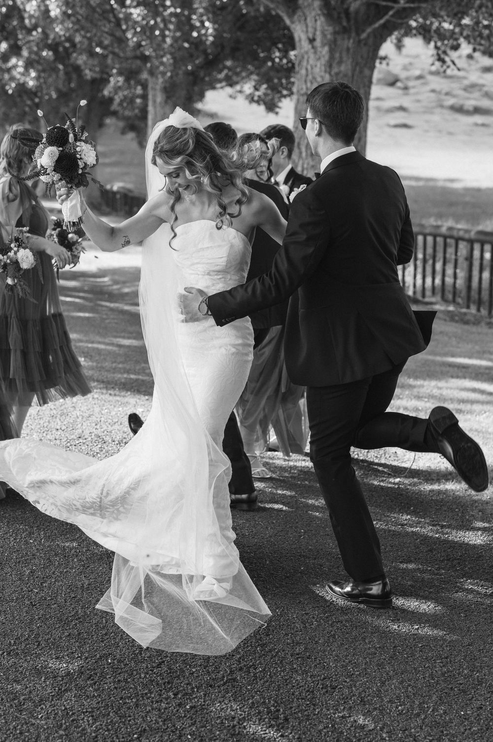 Black and white photo of a bride and groom dancing joyfully outdoors, with wedding party members visible in the background.