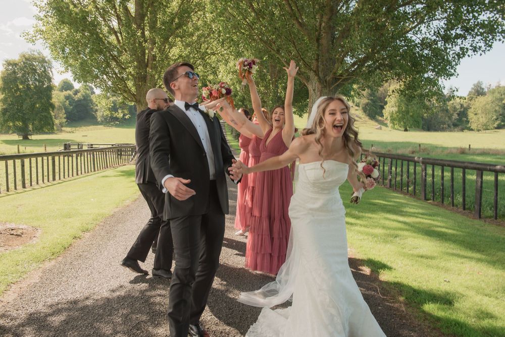 Bride and groom walk joyfully with wedding party on scenic garden path, bridesmaids in pink dresses celebrating outdoors.