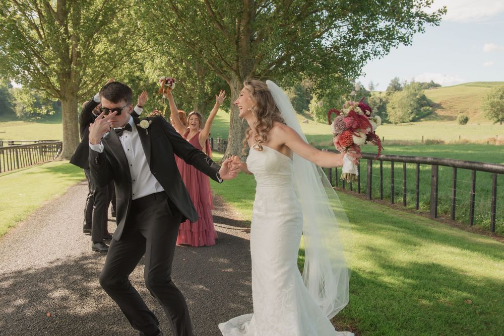 Joyful bride and groom dancing with wedding party on a tree-lined path at an outdoor summer wedding.