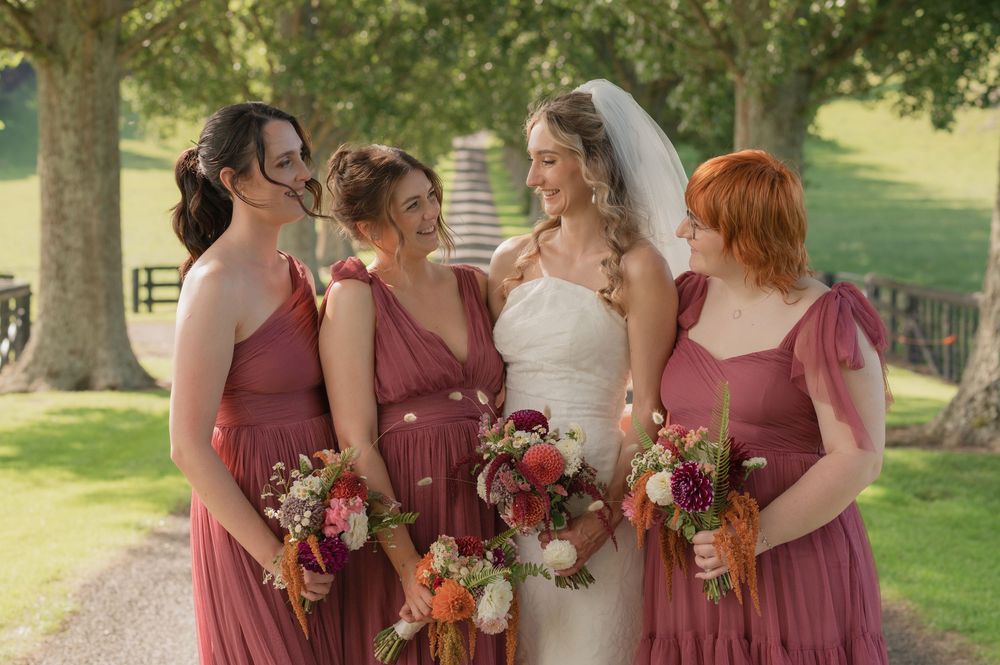 Bride in white gown smiling with three bridesmaids in dusty rose dresses holding colorful bouquets outdoors.