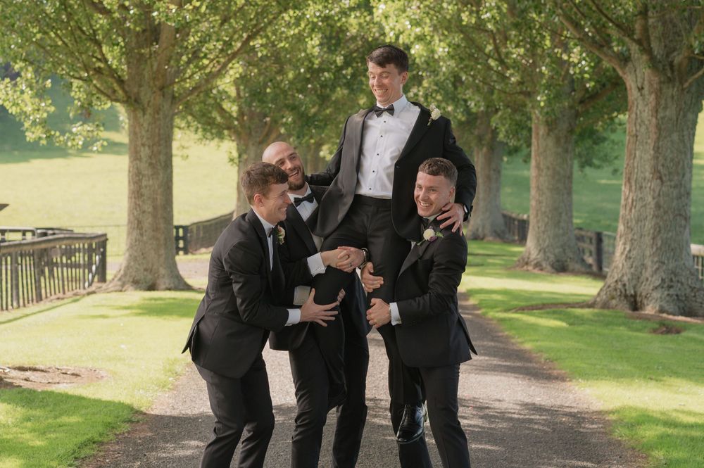Groomsmen in black tuxedos lifting the groom outdoors on a tree-lined path at a wedding celebration.