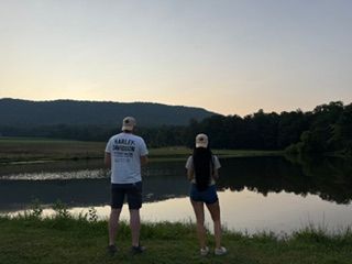 Two people in Harley Davidson shirts stand by a calm reflective pond at sunset with green hills in the background.