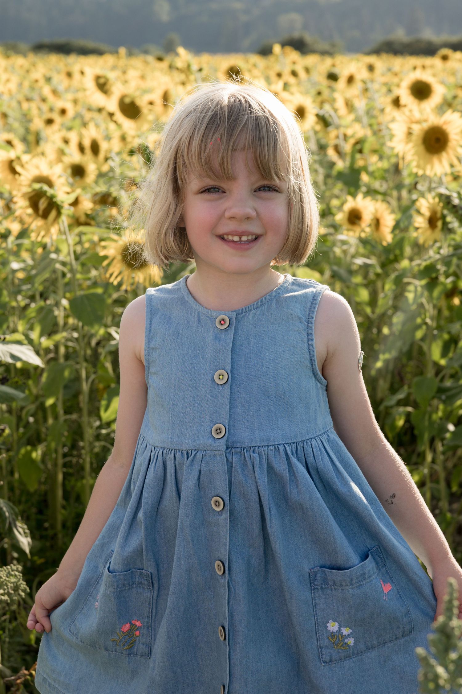 Young blonde girl in denim dress smiling in sunflower field.