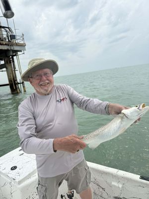 Smiling man holding a large spotted fish on a boat near a pier over calm ocean waters.
