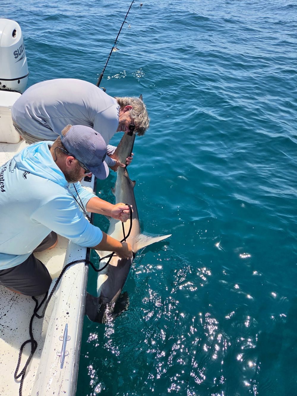 Two fishermen carefully release a shark back into the ocean from their fishing boat.