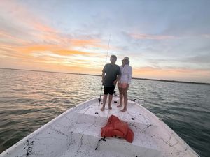 Two people stand on a boat bow fishing at sunset over calm waters with a colorful orange and pink sky.