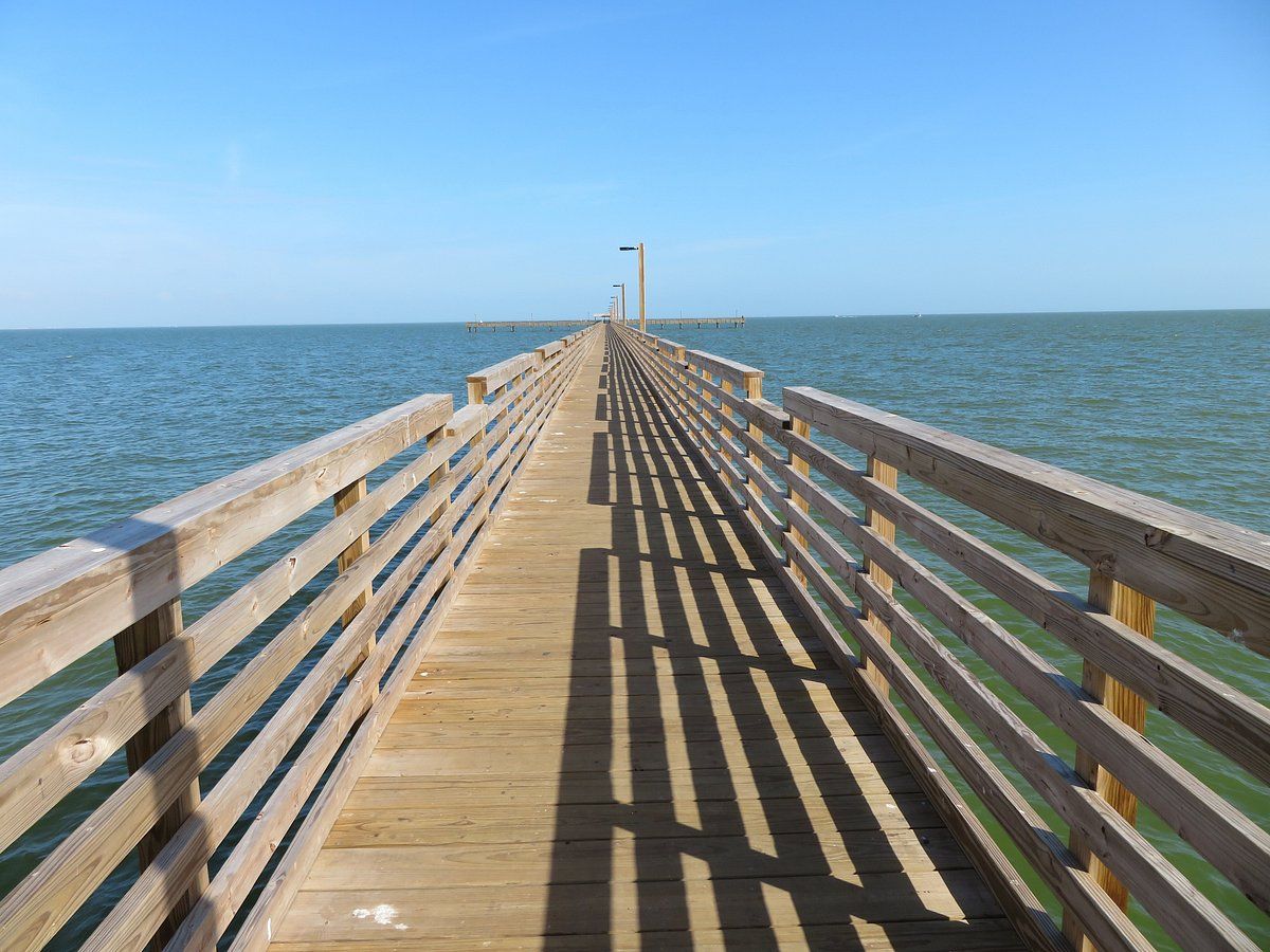 Long wooden fishing pier extending into calm blue coastal waters under a clear sky.