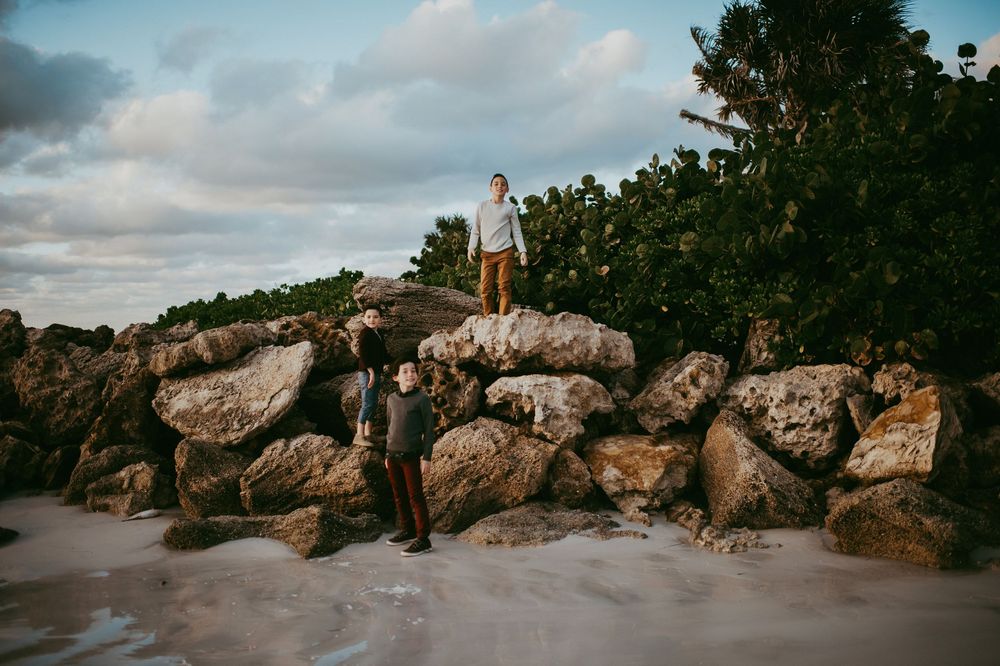 Two men pose on rocky coastal boulders near lush greenery and sandy beach under a cloudy sky.