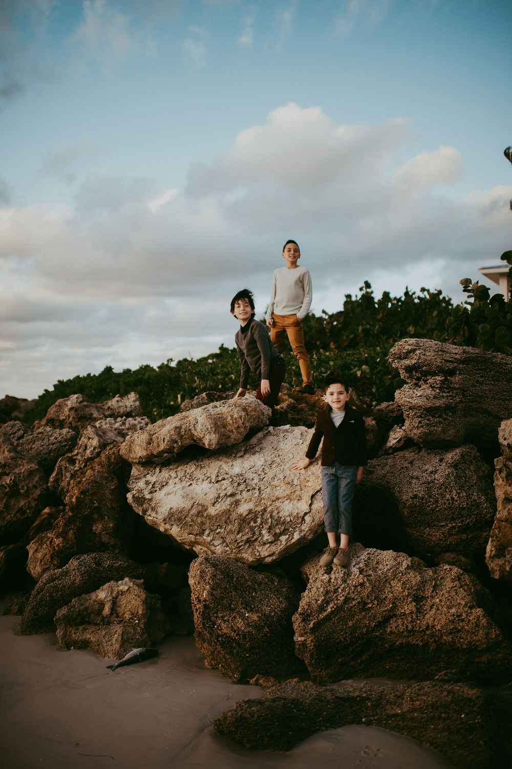 Three people standing on large coastal rocks with greenery and cloudy sky backdrop near a sandy beach.